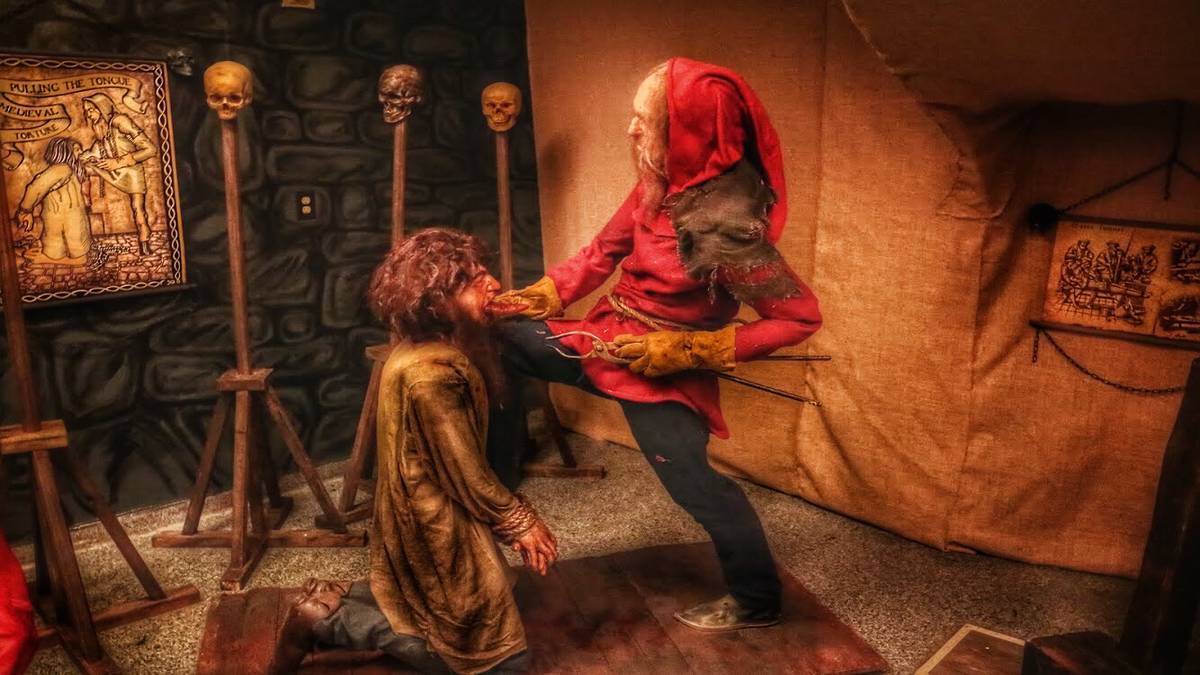 Figurines in a museum, one is kneeling before the other while he holds a clamp and pulls on the kneeling man's tongue